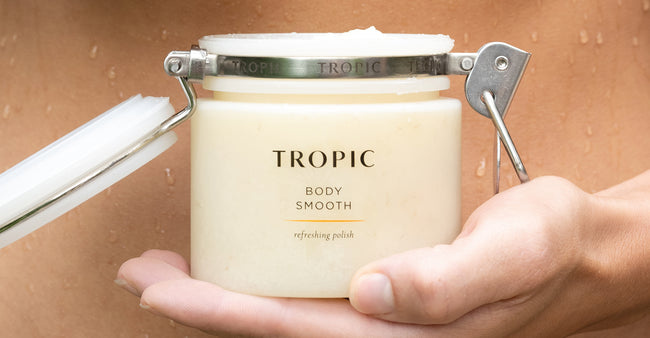 Hand holding an open jar of Tropic “Body Smooth” refreshing polish, with a metal clasp lid and water droplets visible on skin in the background.