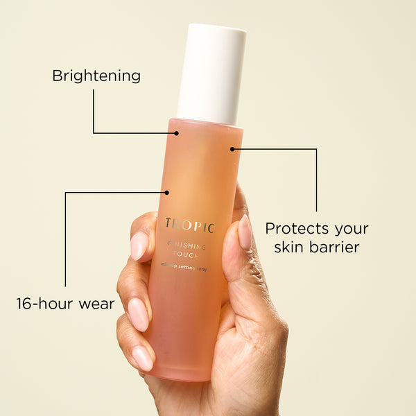 Hand holding a bottle of Tropic Finishing Touch makeup setting spray with text highlighting its benefits: brightening, 16-hour wear, and protection for the skin barrier.