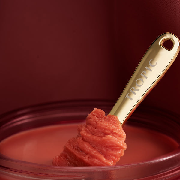 A gold spatula engraved with “TROPIC” scoops a textured, coral-red balm from a glass jar, set against a deep red background.