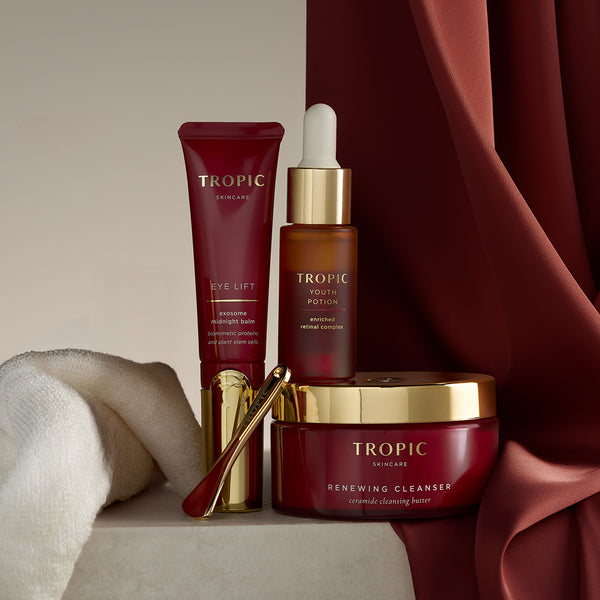 Tropic skincare bundle arranged on a stone surface with rich red fabric, featuring Renewing Cleanser in a red jar with a gold lid, Eye Lift in a red tube with a gold applicator, Youth Potion in a glass dropper bottle, a gold skincare spatula, and a soft bamboo cleansing cloth.