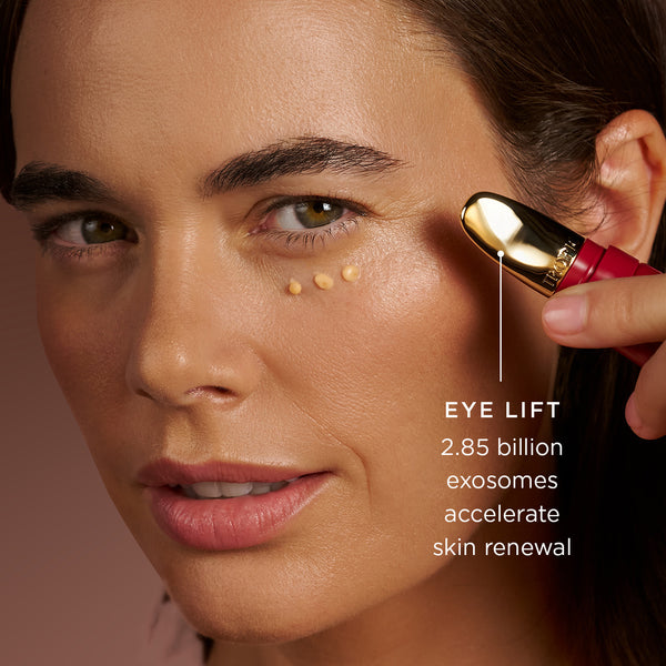Close-up of a model applying Tropic Eye Lift under the eye using a gold applicator, with small dots of product visible on the skin. On image text reads: “Eye Lift – 2.85 billion exosomes accelerate skin renewal”.