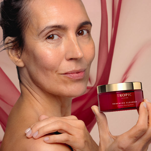 Model holding Tropic Renewing Cleanser in a red jar with a gold lid, shown against a soft pink backdrop.
