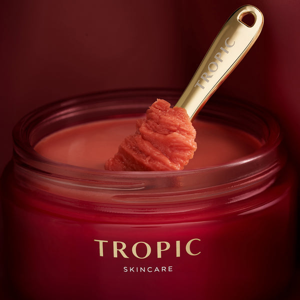 Gold Tropic spatula lifting red toned cleanser from an open deep red Tropic Skincare jar.