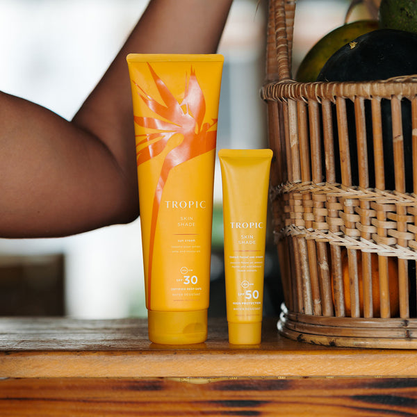 Tropic Skin Shade Sun Cream SPF 30 and Skin Shade Tinted Facial Sun Cream SPF 50 tubes on a wooden surface beside a basket of fruit, offering reef-safe, water-resistant sun protection.