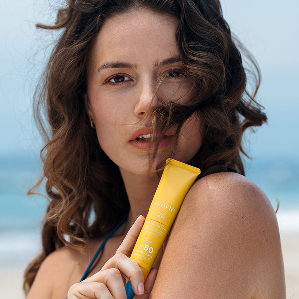Woman with wavy brown hair holding Tropic Skin Shade tinted facial sun cream tube against her shoulder on a sunny beach.