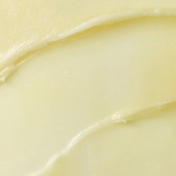 The image is a close-up of a smooth, creamy, pale yellow substance, likely a balm or butter. It shows a glossy texture with gentle curves, suggesting it has been spread or swirled. The rich appearance indicates it might be a hydrating skincare product.