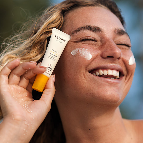 A smiling woman with sun-kissed skin and beachy waves holds a tube of Tropic Sun Care 