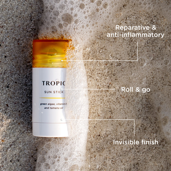 Tropic Sun Stick with green algae, vitamin E, and tamanu oil lying on wet sand with ocean foam, featuring text labels: reparative & anti-inflammatory, roll & go, invisible finish.