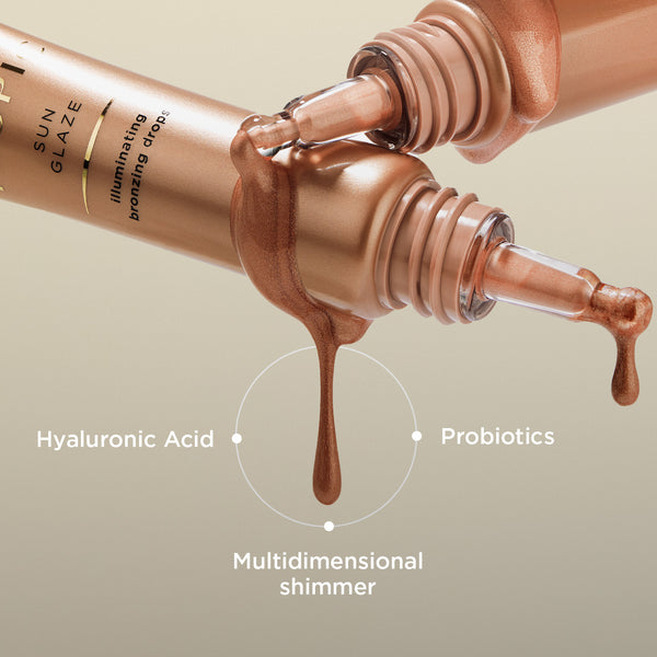 Image shows two Sun Glaze illuminating bronzing drop tubes crossing over one another with the product dripping out of the applicator. The product contains: Hyaluronic Acid, Probiotics and multidimensional shimmer.