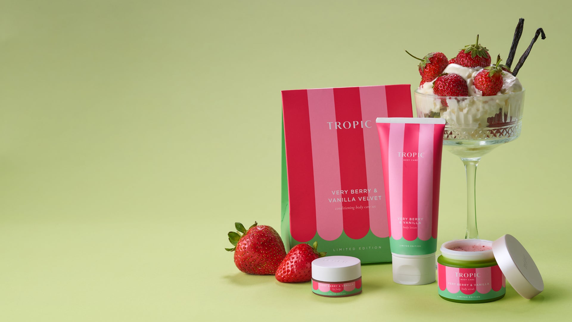 Tropic Body Care’s limited-edition Very Berry & Vanilla Velvet collection displayed on a soft green background. A pink-and-red striped gift box, matching body lotion tube, body scrub jar, and lip fudge pot are arranged with fresh strawberries. Beside the products, a glass dessert dish filled with whipped cream and strawberries, topped with vanilla pods, reinforces the sweet berry-and-vanilla theme. Plenty of empty space appears on the left side of the image.