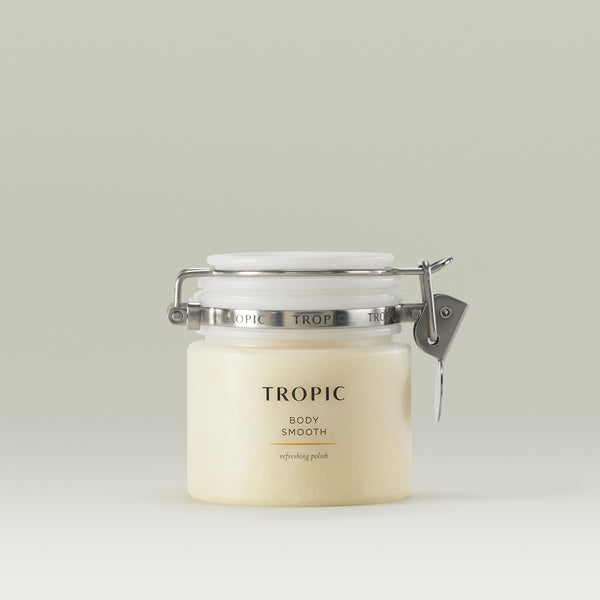 A jar of Tropic Body Smooth refreshing polish, a skincare product in a cream-colored scrub. The jar has a minimalist design with a white lid, metal clasp, and a transparent body showing the product inside. The label features the brand name 'TROPIC' in uppercase letters, followed by 'BODY SMOOTH' and 'refreshing polish' in a smaller font. The background is a soft, neutral shade.
