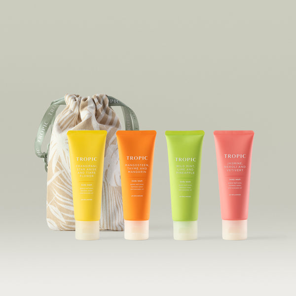 Four Tropic body wash tubes in yellow, orange, green, and coral, displayed in front of a beige drawstring bag with a leaf pattern. The scents shown are Frangipani, Star Anise and Tiare Flower; Mangosteen, Thyme and Mandarin; Wild Mint, Lime and Pineapple; and Jasmine, Neroli and Vetivert.
