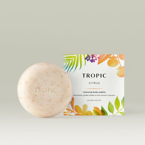 Tropic Citrus Cleansing Body Pebble – a round, exfoliating soap bar with natural flecks, embossed with the Tropic logo, alongside its vibrant tropical-themed packaging. Made with shea butter, golden jojoba oil, and coconut cleansers for a pH-balanced cleanse.