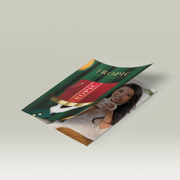 Tropic Luxury Gift Guide 2025 brochure lying partly open on a white surface, featuring the red Tropic gift case on the cover and an inside page showing Susie Ma smiling holding a mug.