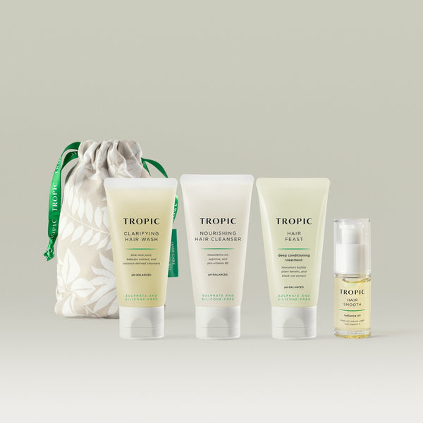 Four Tropic haircare products are arranged on a neutral background beside a cream drawstring pouch with green Tropic ribbon. From left to right: Clarifying Hair Wash, Nourishing Hair Cleanser, Hair Feast deep conditioning treatment, and Hair Smooth radiance oil in a pump bottle.