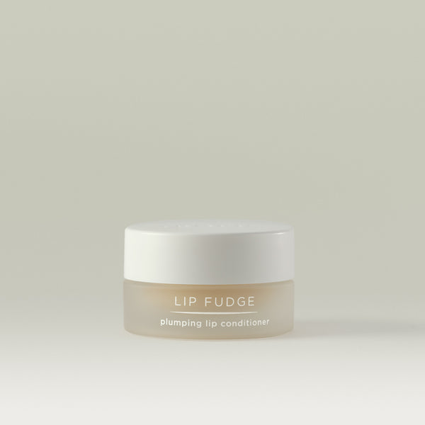 Lip Fudge plumping lip conditioner. Round glass tub with white screw top lid showing Clear's neutral shade through the container.