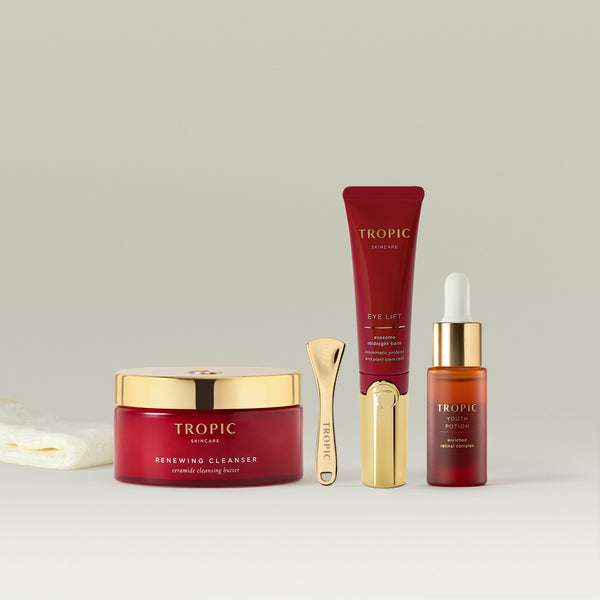 Tropic skincare collection arranged on a soft neutral background, featuring Renewing Cleanser in a red jar with a gold lid, a gold skincare spatula, Eye Lift in a red tube with a gold applicator, Youth Potion in a glass dropper bottle, and a folded bamboo cleansing cloth.