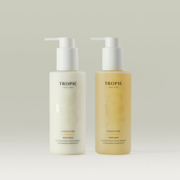 Two bottles of Tropic Body Care Signature hand care products, featuring a hand lotion and a hand wash. Both bottles have a minimalist design with white pump dispensers and subtle floral patterns on the semi-transparent packaging. The hand lotion is in a white bottle, while the hand wash is in a light amber-colored bottle. The labels display 'TROPIC BODY CARE' at the top, followed by 'SIGNATURE' and product details, mentioning Australian lemon myrtle, bergamot, and eucalyptus essential oils. The background i