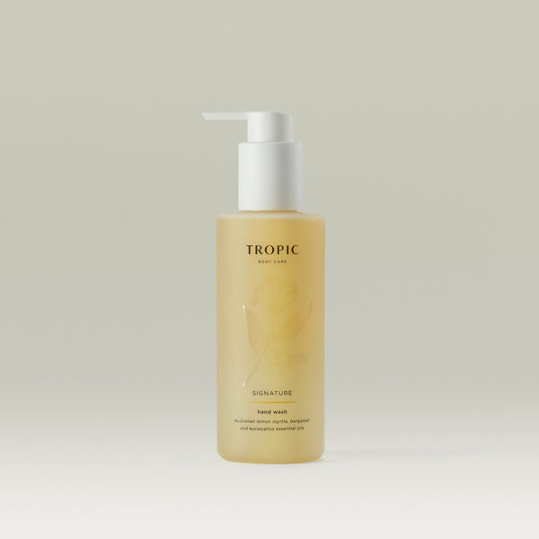 A bottle of Tropic Body Care Signature hand wash with a minimalist design. The semi-transparent, amber-colored bottle features a white pump dispenser and a subtle floral pattern on the packaging. The label displays 'TROPIC BODY CARE' at the top, followed by 'SIGNATURE' and 'hand wash.' The product contains Australian lemon myrtle, bergamot, and eucalyptus essential oils. The background is a soft, neutral shade.