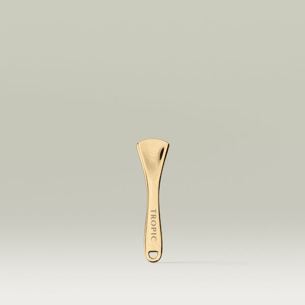 Gold Tropic Skincare spatula standing upright against a soft neutral background, with the word “TROPIC” engraved on the handle.