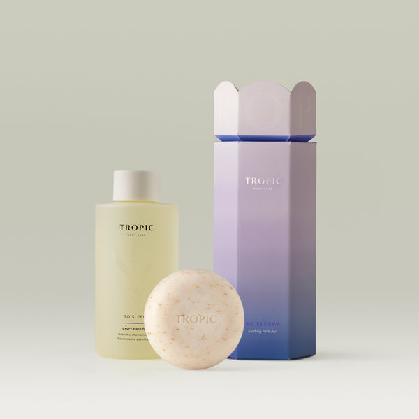 Tropic Body Care So Sleepy bath duo featuring a pale yellow bath foam bottle, a speckled cleansing body pebble, and a lavender-to-blue gradient gift box, styled on a soft neutral background.