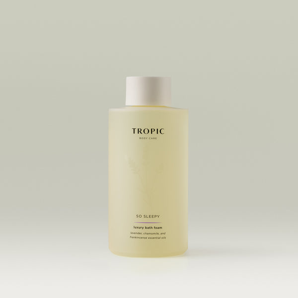A bottle of Tropic So Sleepy Luxury Bath Foam, a pale yellow liquid in a frosted cylindrical bottle with a white cap. The label reads ‘Tropic Body Care – So Sleepy – luxury bath foam with lavender, chamomile, and frankincense essential oils,’ placed against a neutral light background.