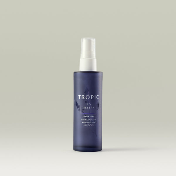 Bottle of Tropic So Sleepy Pillow Mist, the bottle is a deep purple color and features a spray nozzle. The mist contains lavender, chamomile, and frankincense essential oils. The product name and ingredients are displayed in white lettering, and the bottle has a minimalistic design with subtle botanical graphics.