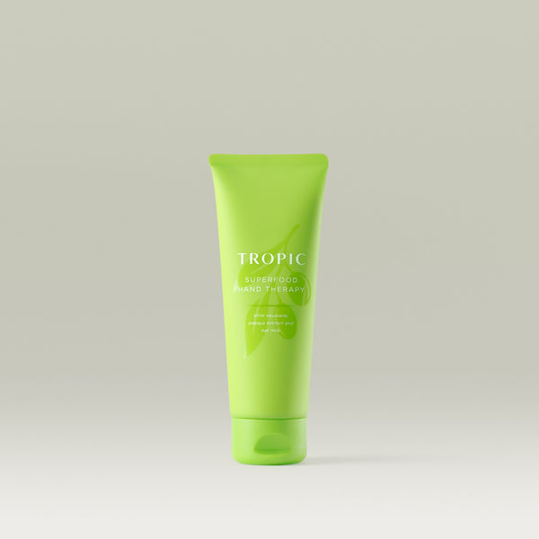 The image shows a green tube of Tropic Superfood Hand Therapy featuring ingredients like olive squalane, papaya extract, and oat milk.