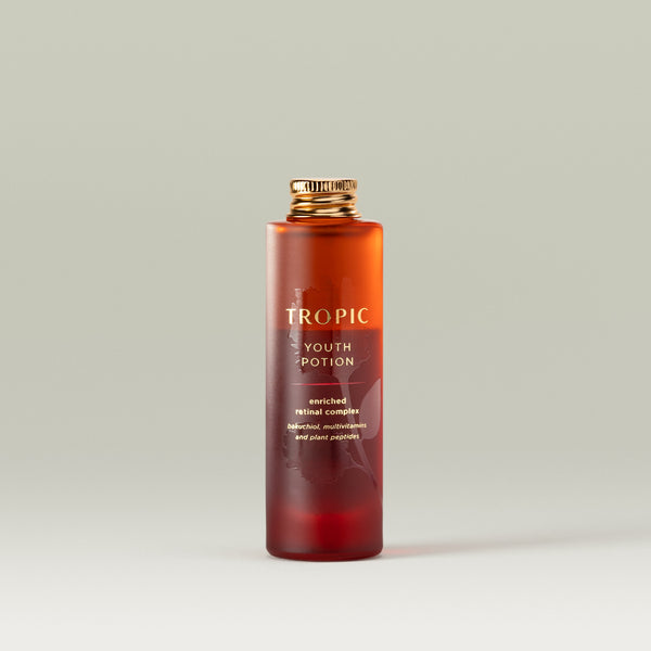 An open frosted amber glass bottle of Tropic Youth Potion skincare serum without its cap, standing upright against a soft pale green background. The label reads “Tropic Youth Potion – enriched retinal complex,” with smaller text mentioning bakuchiol, multivitamins, and plant peptides.