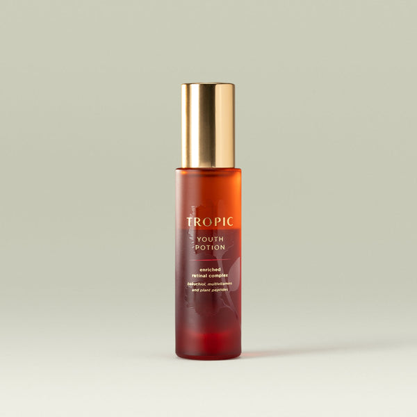 A frosted amber glass bottle of Tropic Youth Potion skincare serum with a gold cap, standing upright against a soft pale green background. The label reads “Tropic Youth Potion – enriched retinal complex” with smaller text mentioning bakuchiol, multivitamins, and plant peptides.