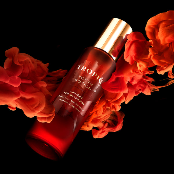 A red Tropic Skincare Youth Potion bottle with a gold lid is angled against a black background, surrounded by flowing, smoke-like swirls of red and orange liquid, creating a dramatic, glowing effect around the product.