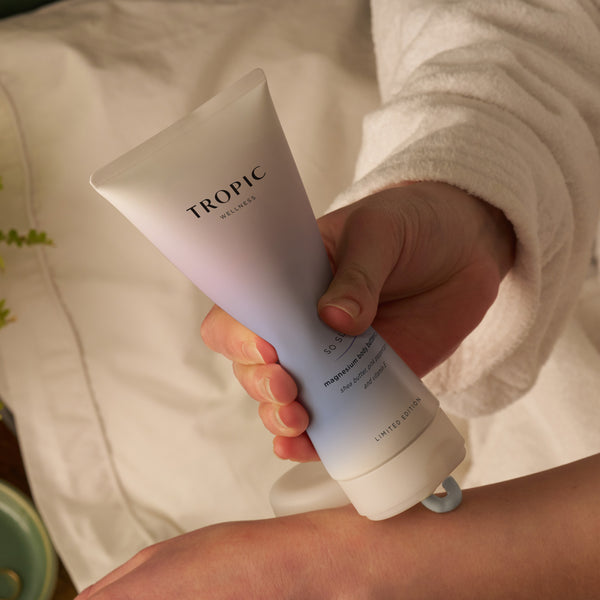 Close-up of a person in a white robe applying Tropic So Sleepy magnesium body butter to their arm from a pastel blue tube.